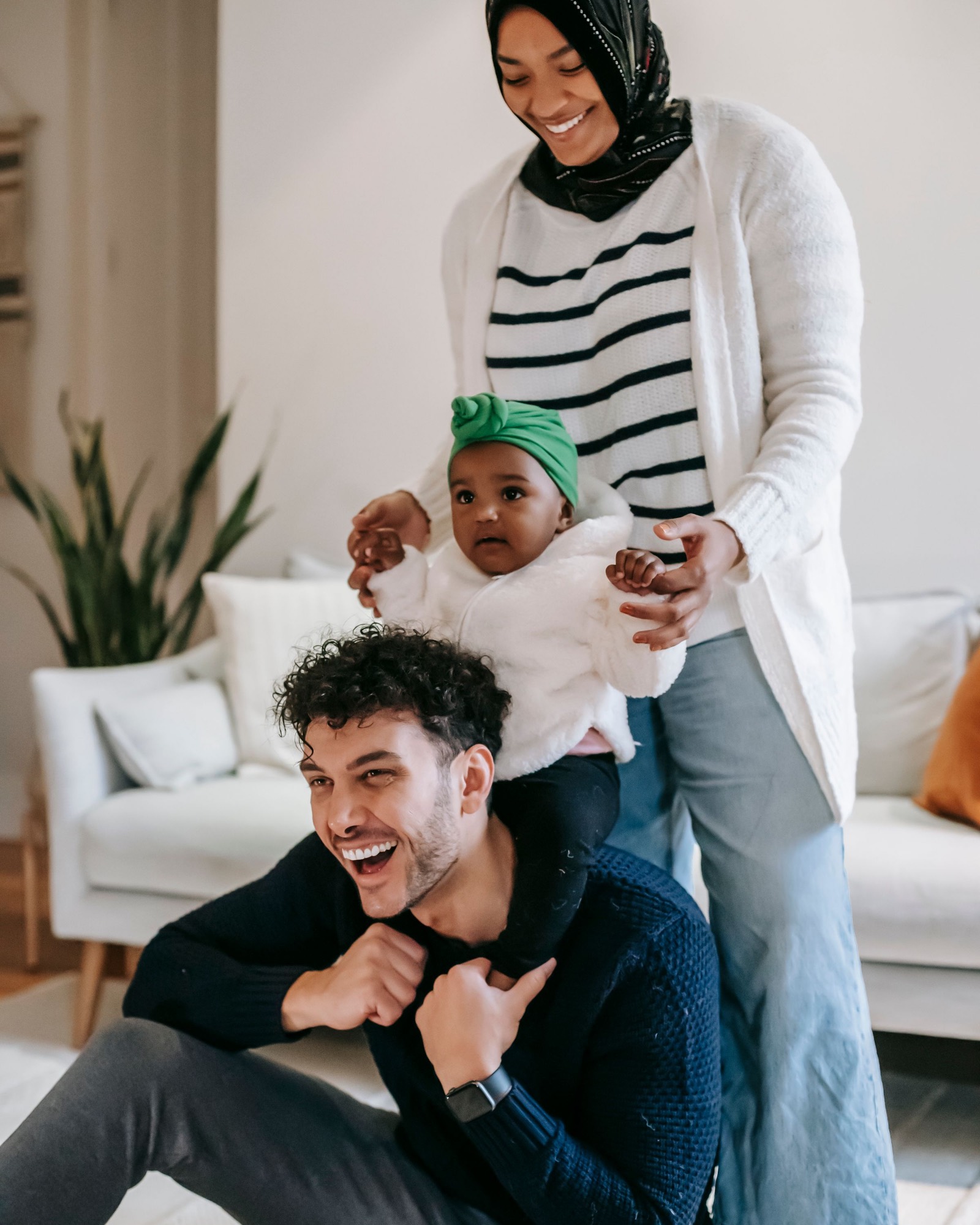 A man laughing with a baby on his shoulders while a woman stands beside them in a bright living room.