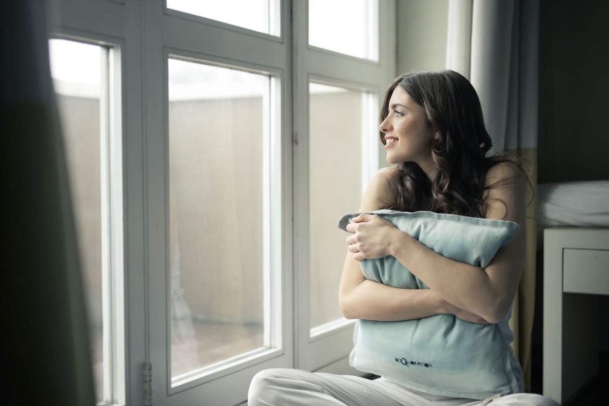 A woman sitting by a window at home, holding a cushion and looking outside.