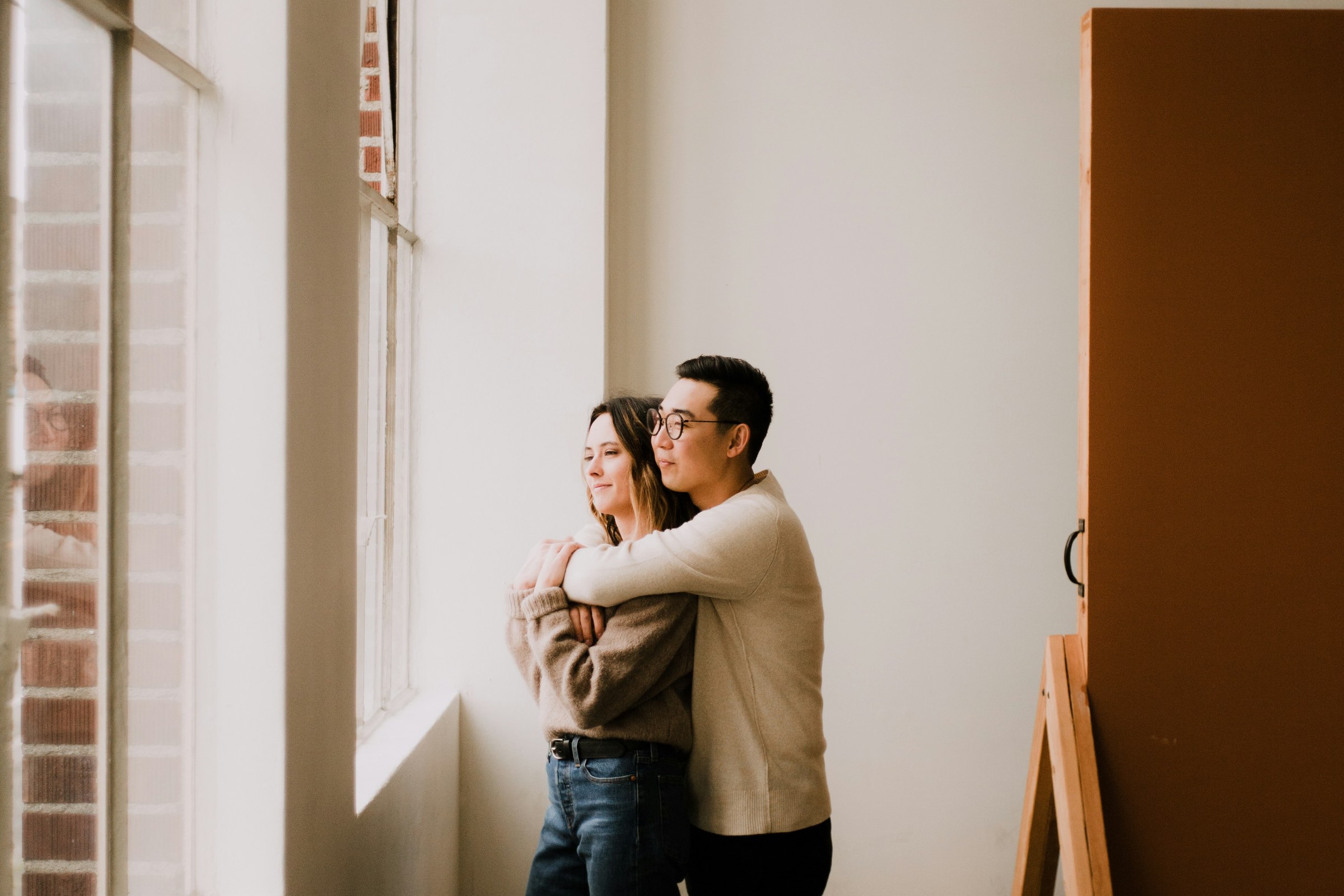 A couple standing together beside tall windows in a bright, sparsely furnished room.