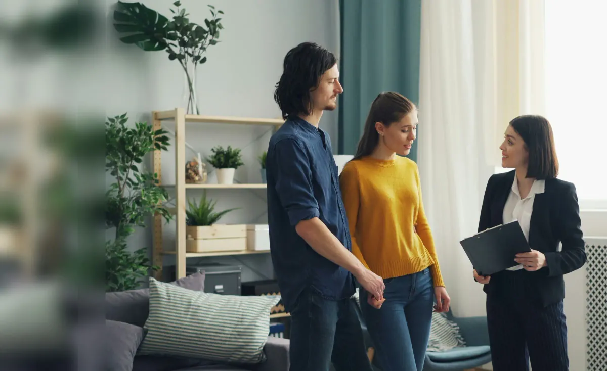 An agent with a clipboard talking with a young couple in the bright living room of a home they are viewing.