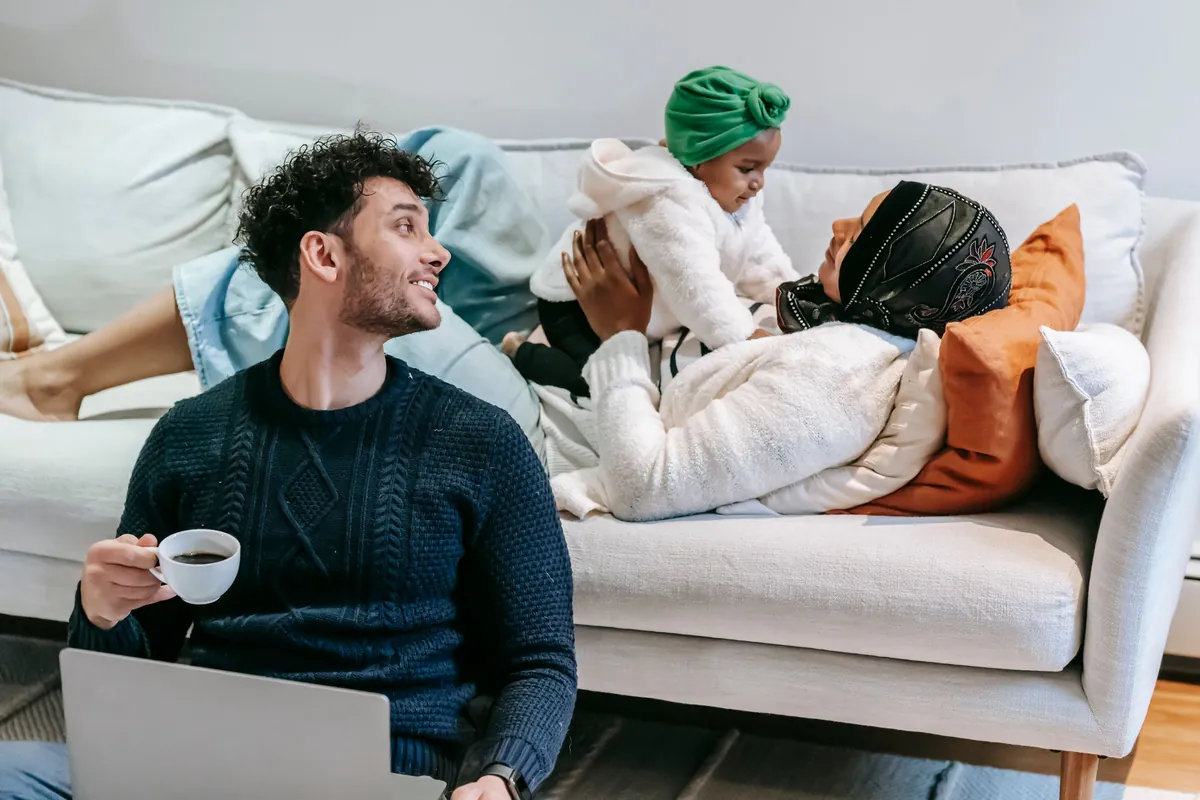 A woman lying on a sofa lifts her baby up while a man sitting on the floor with a coffee turns to smile at them.