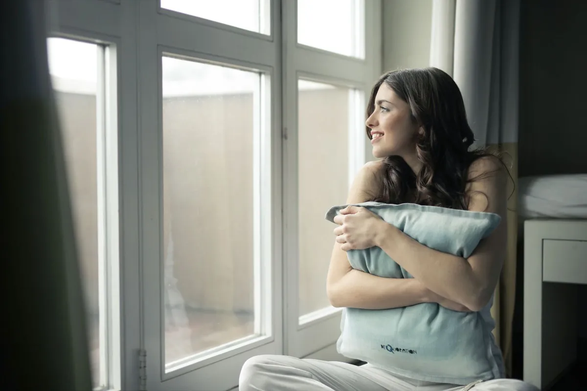 A woman sitting by a window at home, holding a cushion and looking outside.